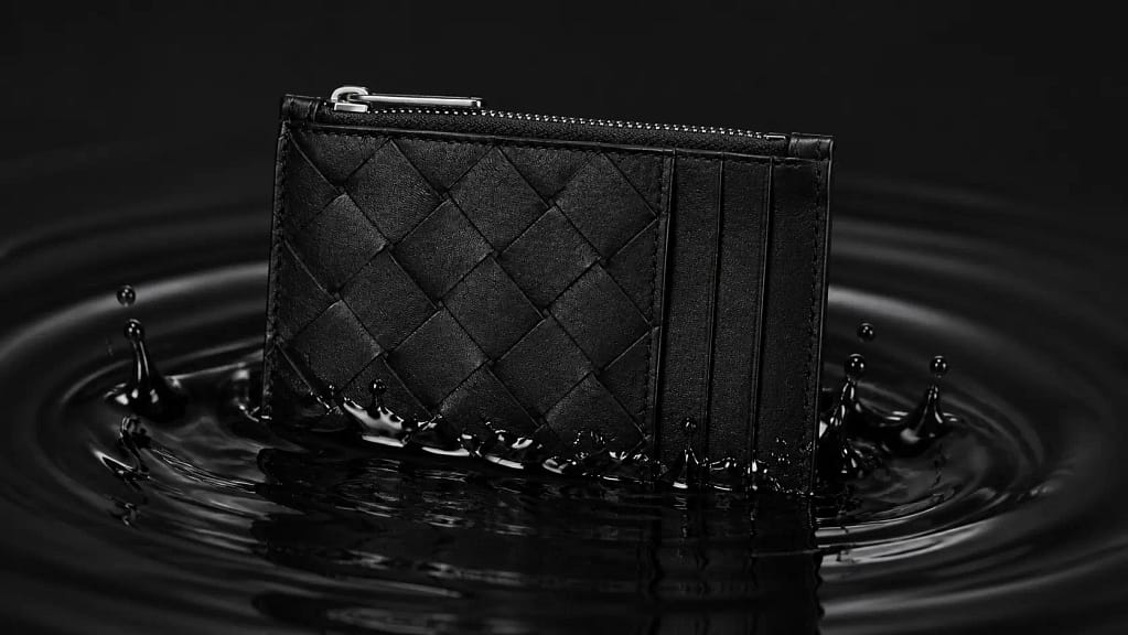 Intrecciato Leather Card Holder encased in a clear ice block, showcasing its black woven texture and silver zipper against a dark reflective surface.